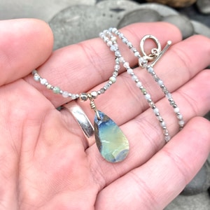 May include: A delicate necklace featuring a teardrop-shaped pendant with blue and yellow hues. The pendant hangs from a beaded chain with a silver clasp. The necklace is displayed on a hand.