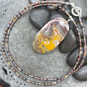 May include: A necklace with a large, oval-shaped agate pendant with brown, yellow, and white swirling patterns. The necklace is made of small, silver, brown, and gold beads.