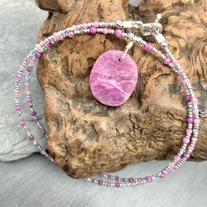 May include: A layered necklace featuring a large, oval-shaped pink pendant. The pendant is suspended from a double strand of small pink and silver beads. The necklace is displayed on a piece of textured wood.