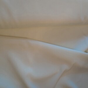 May include: A close-up of a smooth, off-white fabric with a soft, drapey texture. The fabric is folded in half, creating a subtle crease.
