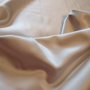 May include: A smooth, silky fabric in a light beige color. The fabric is draped and folded, showcasing its luxurious texture.
