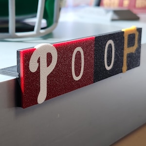 May include: A rectangular sign with the word "POOP" in white, stylized lettering. The sign is divided into three sections: red, black, and black with yellow accents. The sign is resting on a white surface.