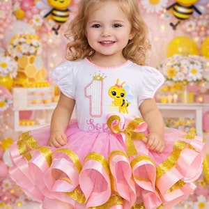 May include: A young child wearing a white t-shirt with a bee graphic and a pink tutu with gold trim. The shirt has the number 1 and a crown. The child is smiling, with a yellow bow in their hair. The background includes balloons and decorations.