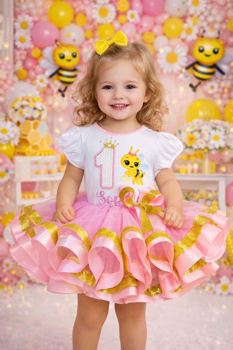 May include: A young child wearing a white t-shirt with a bee graphic and a pink tutu with gold trim. The shirt has the number 1 and a crown. The child is smiling, with a yellow bow in their hair. The background includes balloons and decorations.