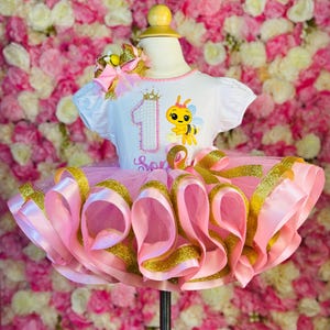 May include: A pink and gold tutu dress with a white top featuring a bee graphic and the number one. The dress has a layered tulle skirt with pink and gold ribbons. A matching bow is attached to the top.