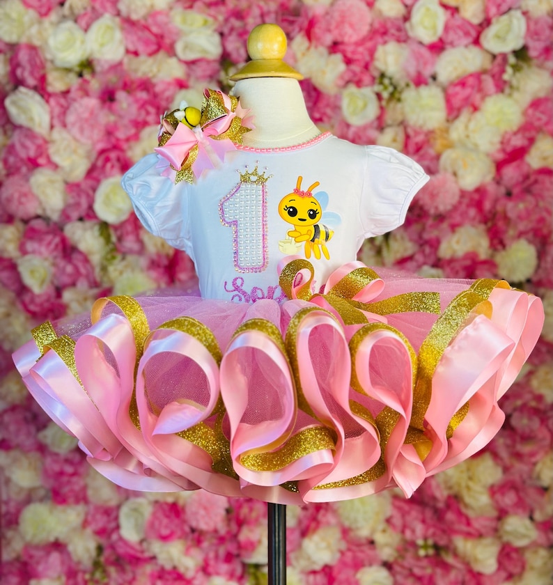 May include: A pink and gold tutu dress with a white top featuring a bee graphic and the number one. The dress has a layered tulle skirt with pink and gold ribbons. A matching bow is attached to the top.