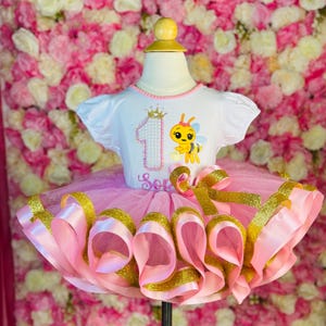 May include: A white short-sleeved top with a pink and gold tutu. The top features a gold crown and the number 1, along with a cartoon bee. The tutu has pink and gold ribbons and a large bow. The name "Sop" is written on the top.