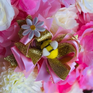 May include: A pink and gold hair bow with a daisy and bee embellishment. The bow features layers of pink satin and gold glitter ribbon. A small, light blue daisy with a yellow center sits atop the bow, and a yellow bee with white wings is attached.