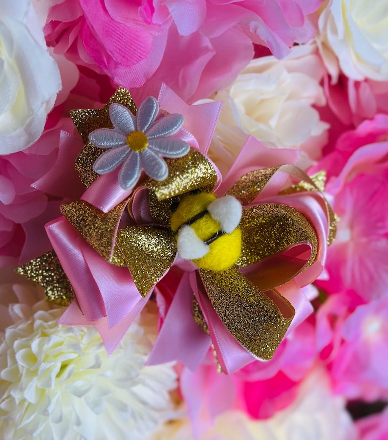 May include: A pink and gold hair bow with a daisy and bee embellishment. The bow features layers of pink satin and gold glitter ribbon. A small, light blue daisy with a yellow center sits atop the bow, and a yellow bee with white wings is attached.