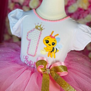 May include: A white toddler's outfit with a pink tutu skirt. The top features a cartoon bee, the number 1, and the name "Serenity". A large gold and pink bow adorns the waist. The outfit is designed for a first birthday.