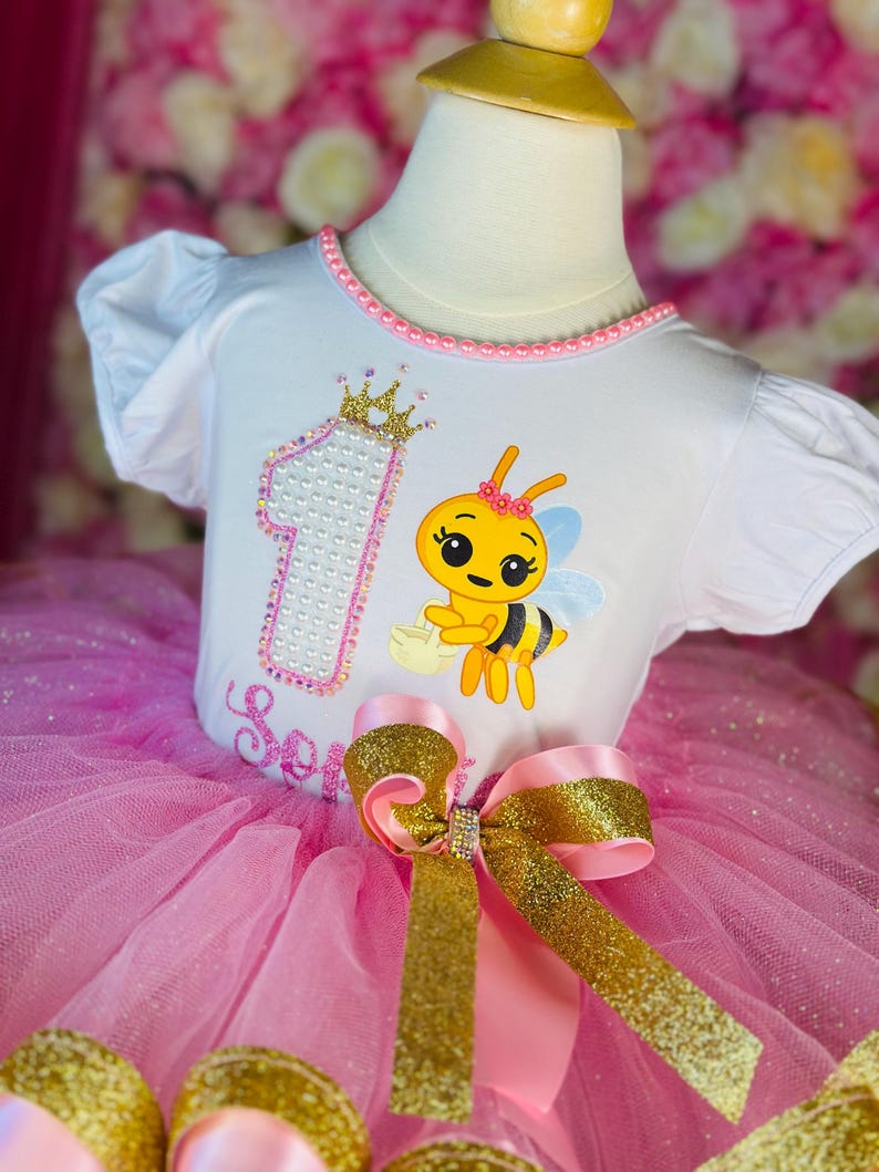 May include: A white toddler's outfit with a pink tutu skirt. The top features a cartoon bee, the number 1, and the name "Serenity". A large gold and pink bow adorns the waist. The outfit is designed for a first birthday.