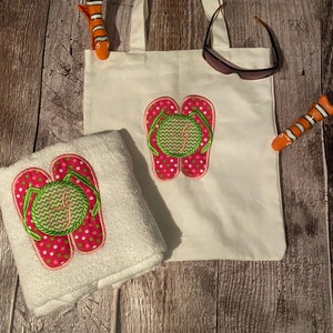 May include: A white canvas tote bag with a pink and green embroidered flip flop design. The bag is next to a white towel with the same design.