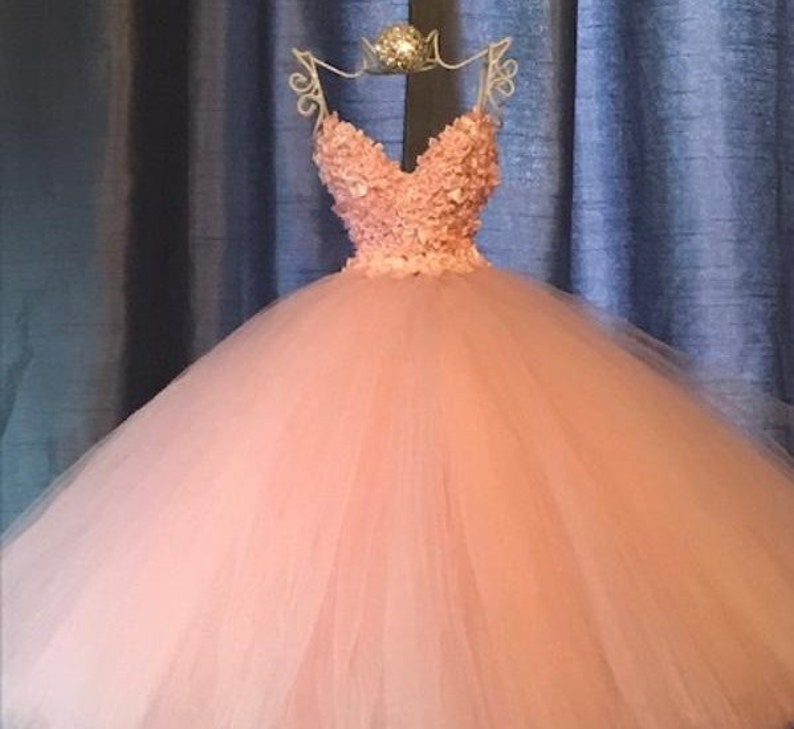 Puede incluir: Un vestido de bola de tul de color melocot&oacute;n con un escote de coraz&oacute;n y un corpi&ntilde;o adornado con delicadas flores rosas.