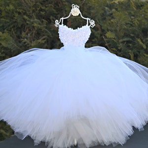 Puede incluir: Un vestido de tul blanco con un corpi&ntilde;o blanco de cuentas. El vestido est&aacute; en una percha blanca y tiene una falda completa.