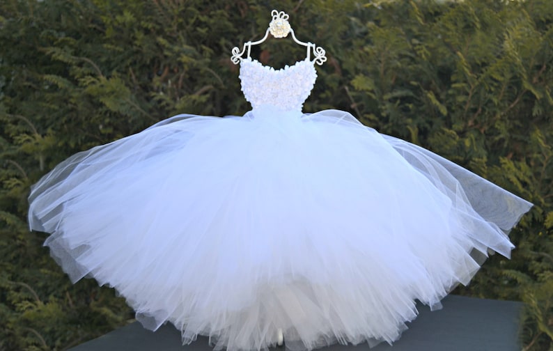 Puede incluir: Un vestido de tul blanco con un corpi&ntilde;o blanco de cuentas. El vestido est&aacute; en una percha blanca y tiene una falda completa.