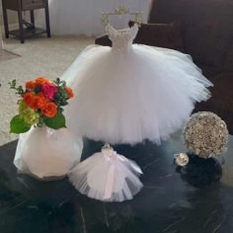 Puede incluir: Un vestido de novia en miniatura de tul blanco con un corpi&ntilde;o de cuentas, un ramo de rosas naranjas, rosas y rojas en un jarr&oacute;n blanco y un peque&ntilde;o vestido de tul blanco con una cinta blanca.