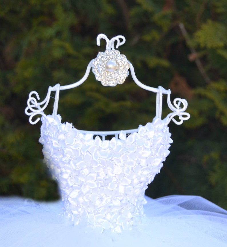 Puede incluir: Tutu de tul blanco con un corpi&ntilde;o de sat&eacute;n blanco decorado con formas de flores blancas. El corpi&ntilde;o est&aacute; en una percha de metal blanco con una flor de pedrer&iacute;a.