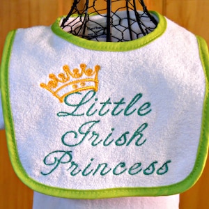 May include: White baby bib with a green trim and a gold crown embroidered on it. The bib says "Little Irish Princess" in green embroidery.