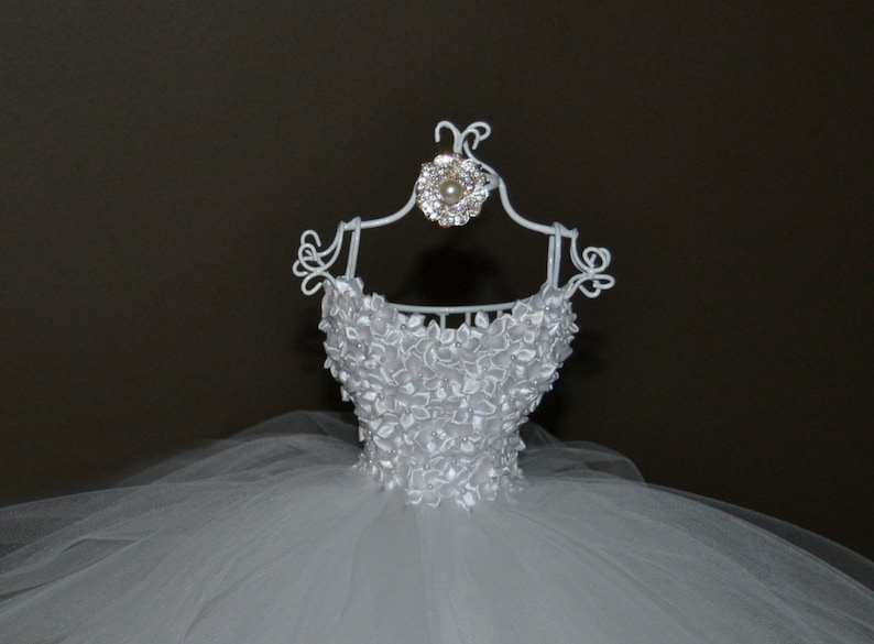 Puede incluir: Vestido de novia de tul blanco en una percha de alambre blanco con un adorno de pedrer&iacute;a y perla.
