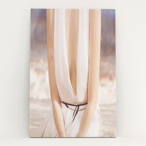 May include: A canvas print featuring a woman in a white dress with a plunging neckline. The dress is cinched at the waist with a black belt. The background has a soft, blurred effect with neutral tones.