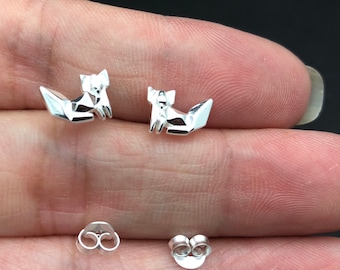 Könnte beinhalten: Ein Paar silberne Fuchs-Ohrstecker. Die Füchse sind stilisiert und geometrisch gestaltet. Die Ohrringe befinden sich auf der Hand einer Person.