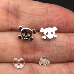 May include: A pair of silver skull earrings with heart-shaped eye sockets. The earrings are on a white background.