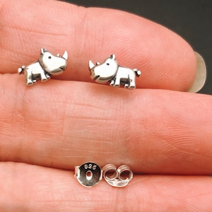 May include: A pair of silver-colored rhino stud earrings. Each earring features a small, detailed rhino design with a horn and small legs. The earrings are shown with their backs and earring posts.