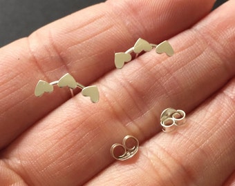 May include: A pair of silver heart shaped earrings with a curved design. The earrings are shown on a hand with the backs of the earrings also visible.
