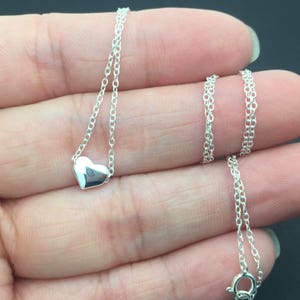 May include: A silver necklace with a small heart pendant. The necklace is on a hand, and the chain is visible.