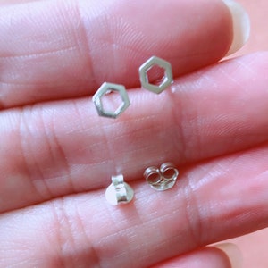 May include: A pair of silver hexagon stud earrings with a simple design. The earrings are shown on a person's hand.