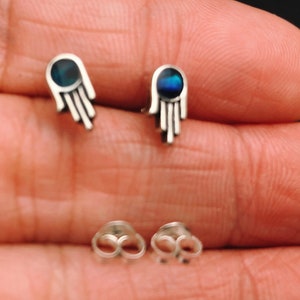 May include: A pair of silver earrings with a hamsa hand design. The earrings feature a blue iridescent stone in the center of each hand.