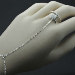 May include: A silver chain bracelet with a heart-shaped charm and a key attached to the charm. The bracelet is connected to a silver ring with a twisted design.