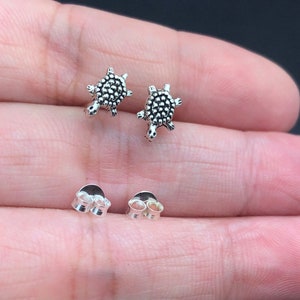 May include: A pair of silver turtle stud earrings with a textured shell design. The earrings are shown on a person's hand.