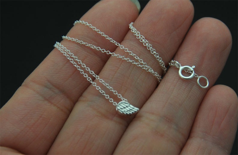 Puede incluir: Un delicado collar de plata con un peque&ntilde;o colgante de ala. El collar tiene una fina cadena y un cierre seguro. El colgante de ala est&aacute; detallado con marcas de plumas individuales. La joya se muestra sobre un fondo oscuro.