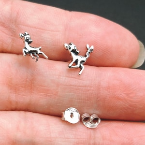 May include: A pair of silver deer stud earrings. The deer are facing different directions and have a detailed design. The earrings are on a person's hand.