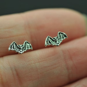 Könnte beinhalten: Ein Paar silberne Fledermaus-Ohrstecker. Die Ohrringe sind klein und zart und haben ein detailliertes Fledermaus-Design.