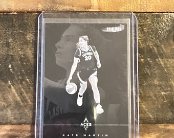 May include: A black and white trading card featuring a basketball player dribbling the ball. The card has a black background with white text that reads "ACES" and "KATE MARTIN". The player is wearing a black and white jersey with the number 20 on the front. The card also has a small white logo with the text "RYAN FORS" in the top right corner.