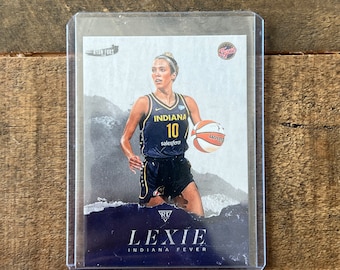 May include: A trading card featuring a basketball player wearing a blue and yellow jersey with the number 10 and the word "INDIANA" on it. The player is holding a basketball and is dribbling the ball. The card has a blue background with the text "LEXIE INDIANA FEVER" on it.
