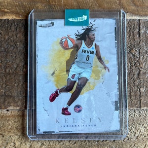 May include: A basketball trading card featuring Kelsey Mitchell of the Indiana Fever. The card is a white background with a yellow and orange watercolor effect. Mitchell is wearing a white jersey with the number 0 and the words "FEVER" and "salesforce".