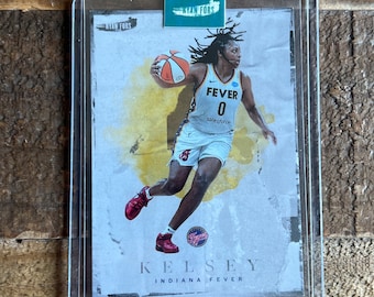 May include: A basketball trading card featuring Kelsey Mitchell of the Indiana Fever. The card is a white background with a yellow and orange watercolour effect. Mitchell is wearing a white jersey with the number 0 and the words "FEVER" and "salesforce".