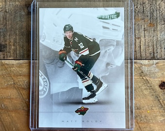 Peut inclure: Carte de hockey représentant Matt Boldy en uniforme des Wild du Minnesota. La carte est dans une pochette de protection, sur fond gris et blanc avec le logo de l'équipe et le nom du joueur.