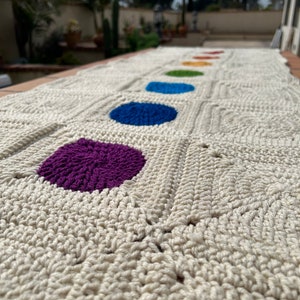 May include: A white crocheted blanket with a row of colorful circles in the center. The circles are purple, blue, green, yellow, orange, and red.
