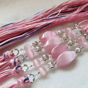 Peut inclure: Gros plan sur des pompons roses et blancs avec des anneaux en métal argenté et des perles de pierres précieuses roses. Les perles sont de forme ovale et entrecoupées de perles plus petites et rondes. Les pompons sont faits de fils roses, blancs et bleus.