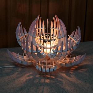 May include: A wooden lotus flower lamp with a warm glow. The lamp is made of light-colored wood and has a detailed, layered design. The petals are a soft pink, and the lamp casts a patterned shadow.