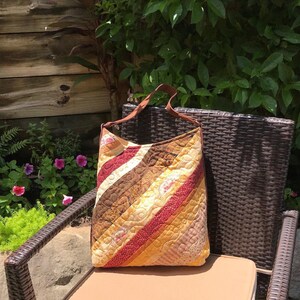 May include: A quilted hobo bag featuring diagonal stripes in shades of brown, tan, orange, and red. The fabric displays a textured, stitched pattern throughout the surface. The bag includes a single brown strap attached to the top corners.