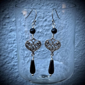 May include: A pair of silver-toned earrings with black accents. Each earring features a black bead, a filigree heart, and a teardrop-shaped black bead. The earrings are suspended from silver hooks.