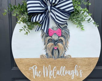 May include: A round wooden door hanger with a white background and a brown wood bottom. A painted image of a Yorkshire Terrier dog with a pink bow is on the white background. The text "The McCullogh's" is written in white on the brown wood bottom. A black and white striped bow with green leaves is on top of the door hanger.