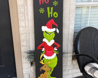 May include: A tall, rectangular black wooden porch sign featuring the text Ho Ho Ho in white, red, and green with green snowflakes. Below, a green character in a red Santa suit stands with a brown dog, followed by the text MERRY GRINCH MAS.