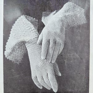 May include: A pair of white, open-weave crochet gloves. The gloves have a delicate, lacy appearance with a scalloped edge on the wrist. The gloves are shown against a dark background.