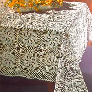Op de afbeelding: Wit gehaakt tafelkleed met een delicaat bloemenpatroon. Het tafelkleed ligt op een houten tafel en heeft een gegolfde rand.